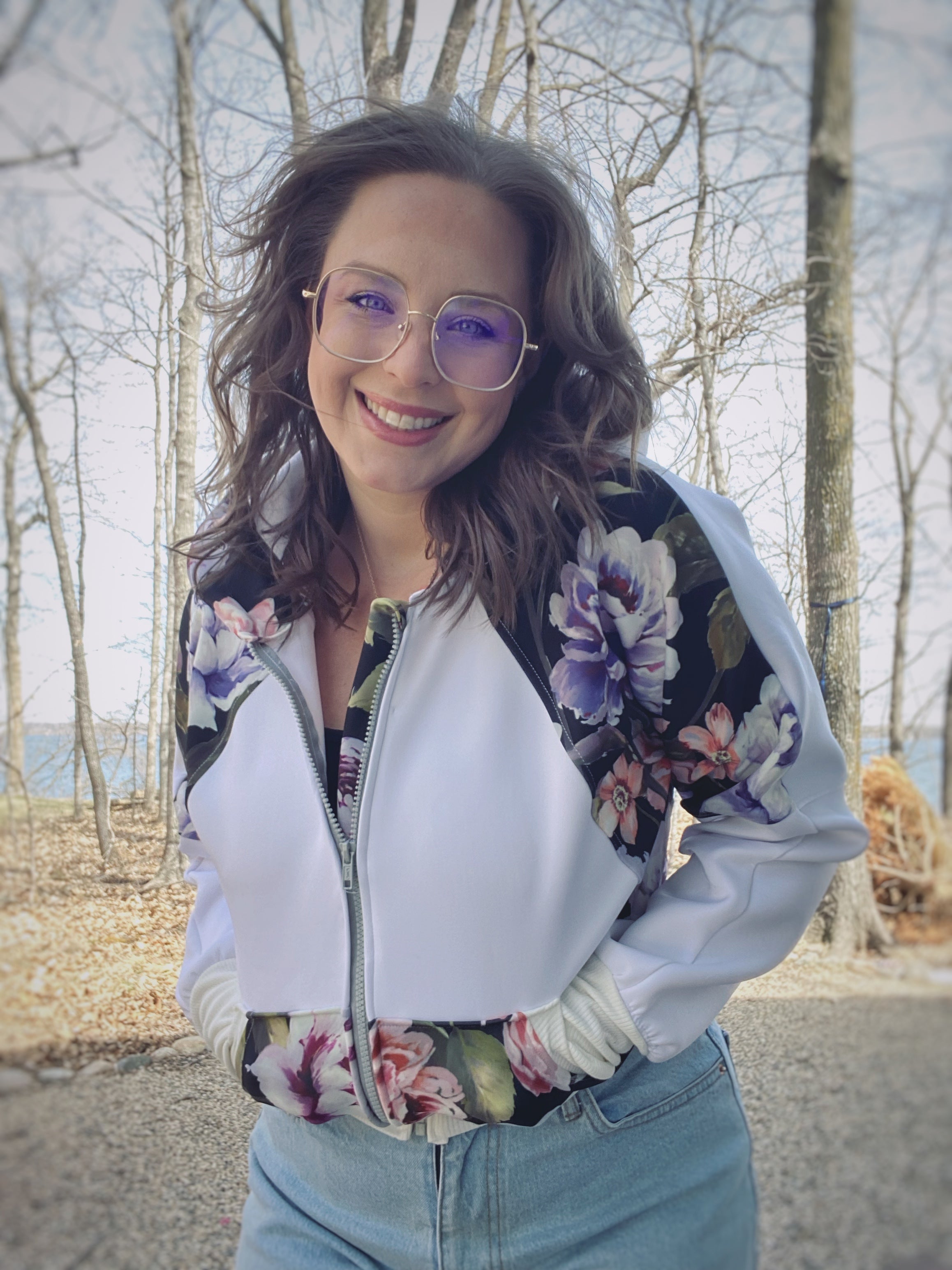 Woman outside by a lake modeling a bright color-block white and floral athletic bomber jacket with zip closure and scuba hood. Cropped version. Ellie and Mac PDF sewing patterns.
