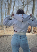 Woman outside by a lake modeling a  color-block white and floral athletic bomber jacket with zip closure and scuba hood. Showing the back. Cropped version. Ellie and Mac PDF sewing patterns.
