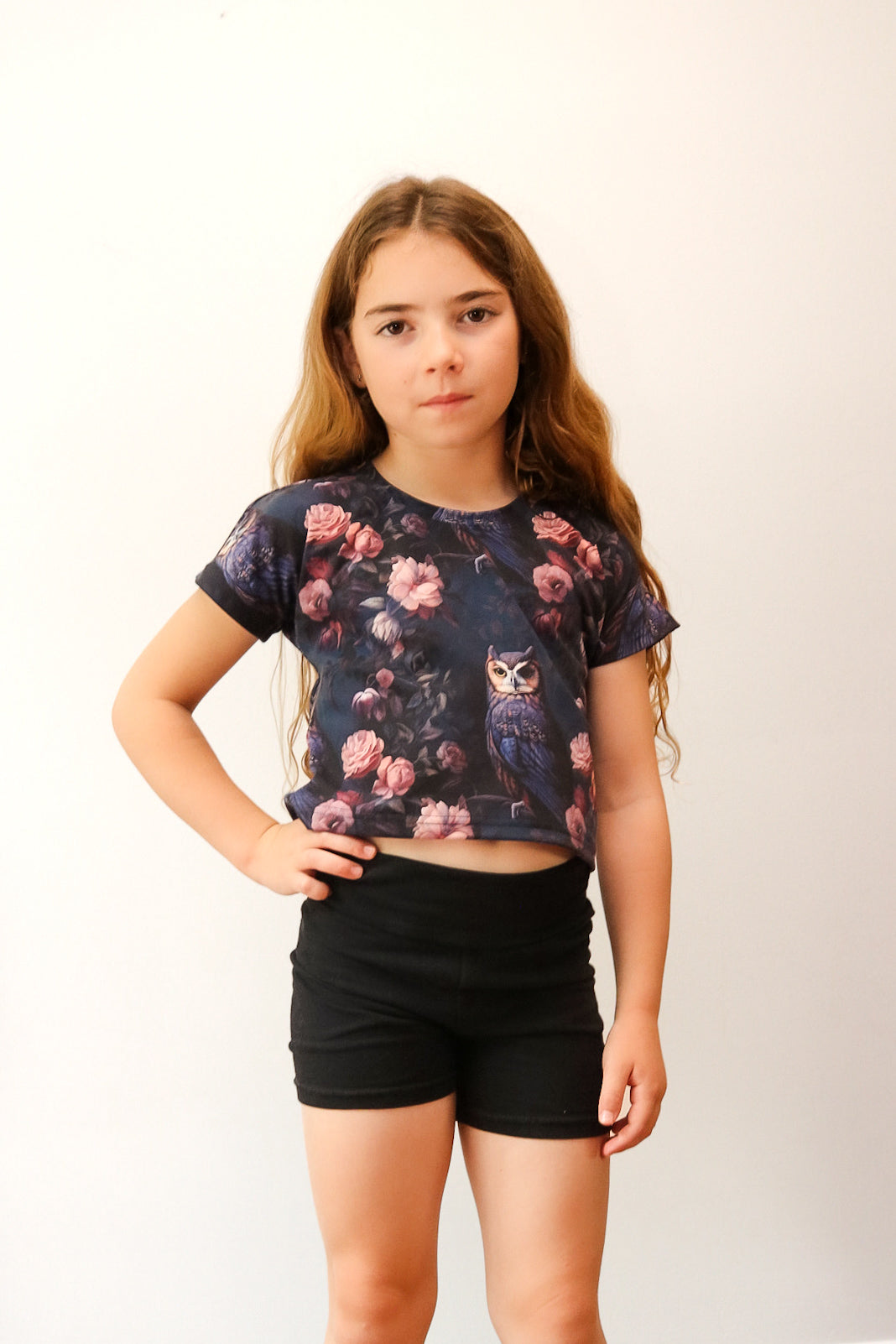 Young girl wearing a floral owl t-shirt and black shorts on a white background