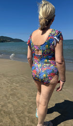 Person wearing a colorful swimsuit on a beach with clear blue sky. The swimsuit was made with the tropical twist swimsuit pdf sewing pattern. It features cut outs in front and back. 