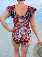 Person wearing a colorful sewing themed fabric swimsuit on a beach. The swimsuit was made with the tropical twist swimsuit pdf sewing pattern. It features cut outs in front and back. 