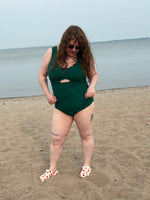 Person wearing a green swimsuit standing on a sandy beach with a gray sky. The swimsuit was made with the tropical twist swimsuit pdf sewing pattern. It features cut outs in front and back. 