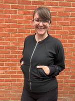 Woman outside by a brick wall modeling a black athletic bomber jacket with zip closure and kangaroo pocket detail. Easy pdf sewing pattern.
