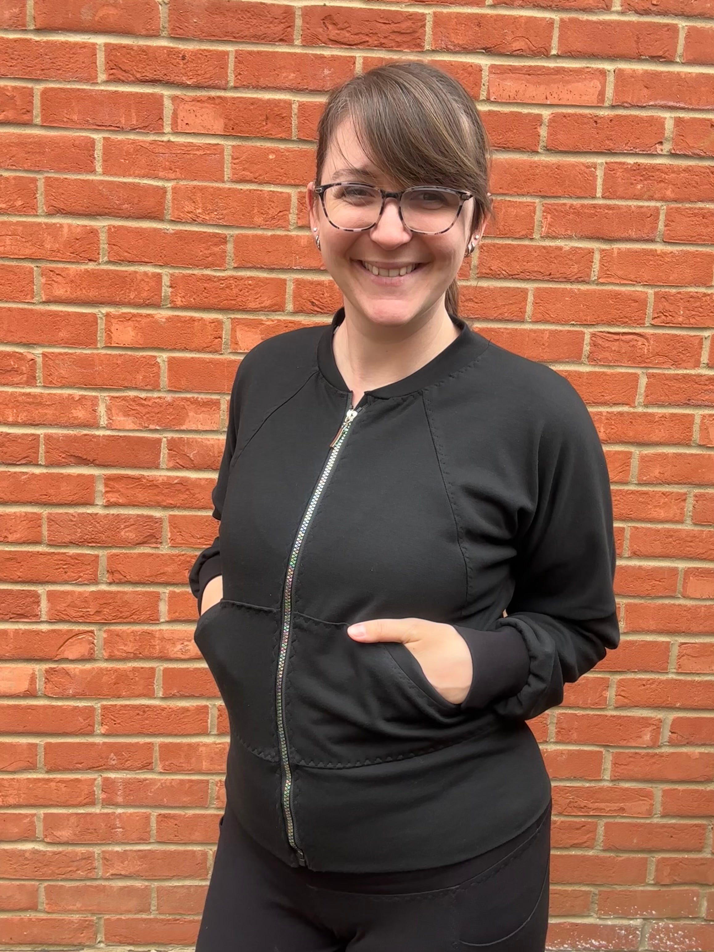 Woman outside by a brick wall modeling a black athletic bomber jacket with zip closure and kangaroo pocket detail. Easy pdf sewing pattern.
