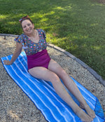 Woman sitting on a blue striped towel in a backyard wearing a burgundy and floral swimsuit. The swimsuit was made with the tropical twist swimsuit pdf sewing pattern. It features cut outs in front and back. 