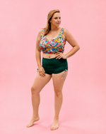 Woman wearing a colorful floral top and green shorts on a pink background. PDF sewing pattern swimwear.