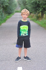 Young boy standing on a path surrounded by greenery wearing a dark blue sweater with a colorful design and black shorts.
