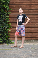 Person wearing a black t-shirt and colorful shorts standing against a wooden wall with greenery.