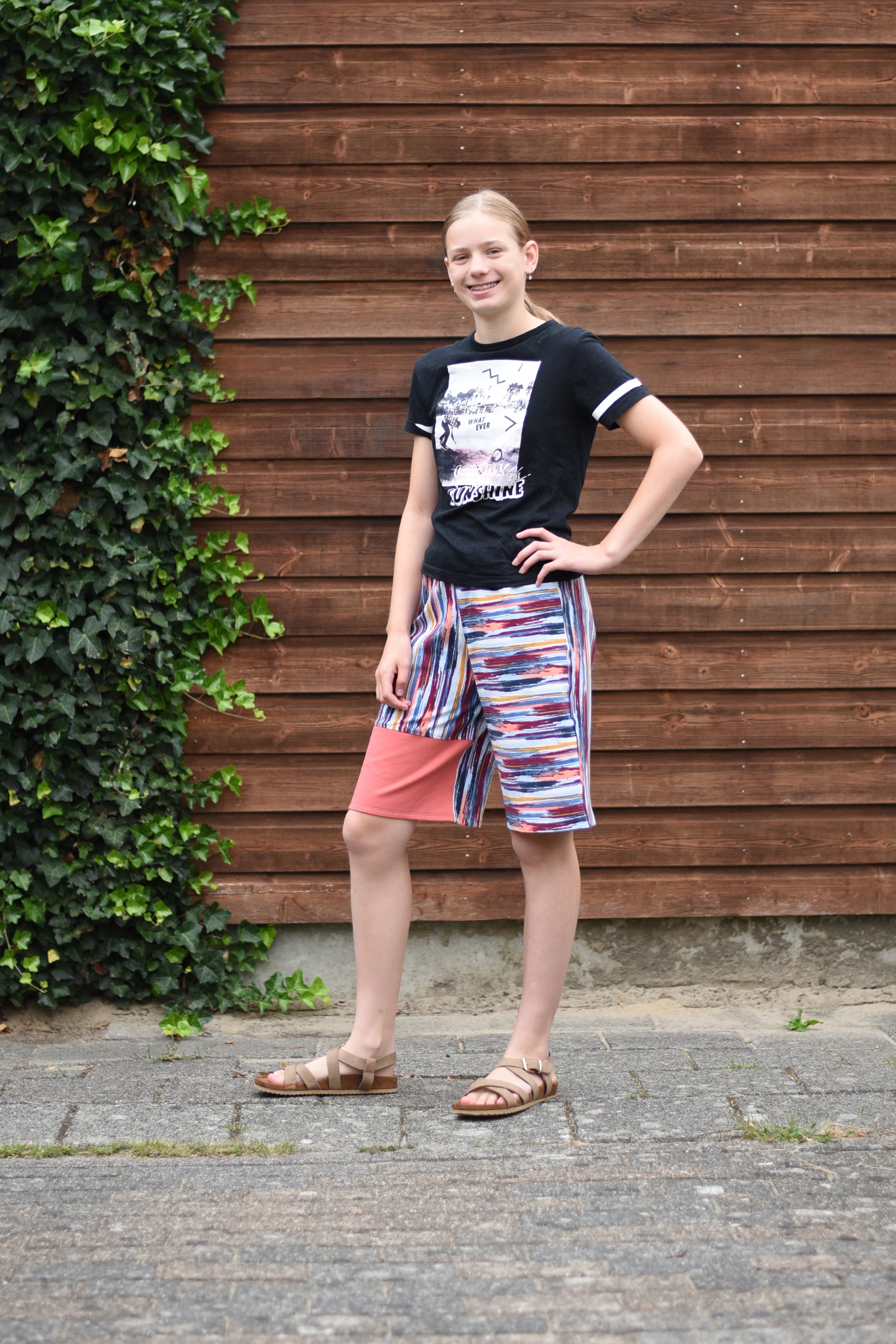 Person wearing a black t-shirt and colorful shorts standing against a wooden wall with greenery.