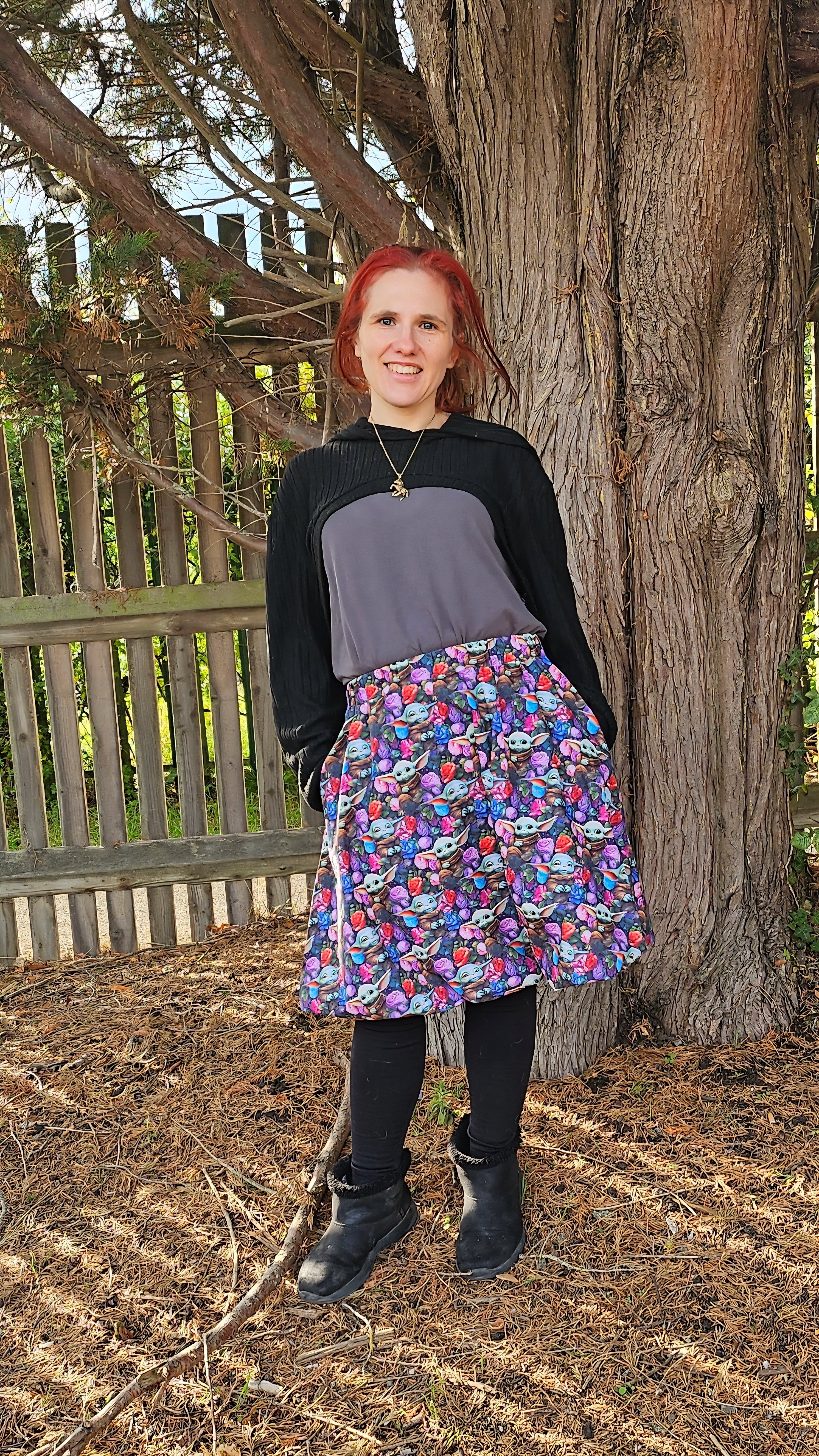 Woman standing outdoors next to a large tree, wearing a colorful character print bubble skirt and black top.