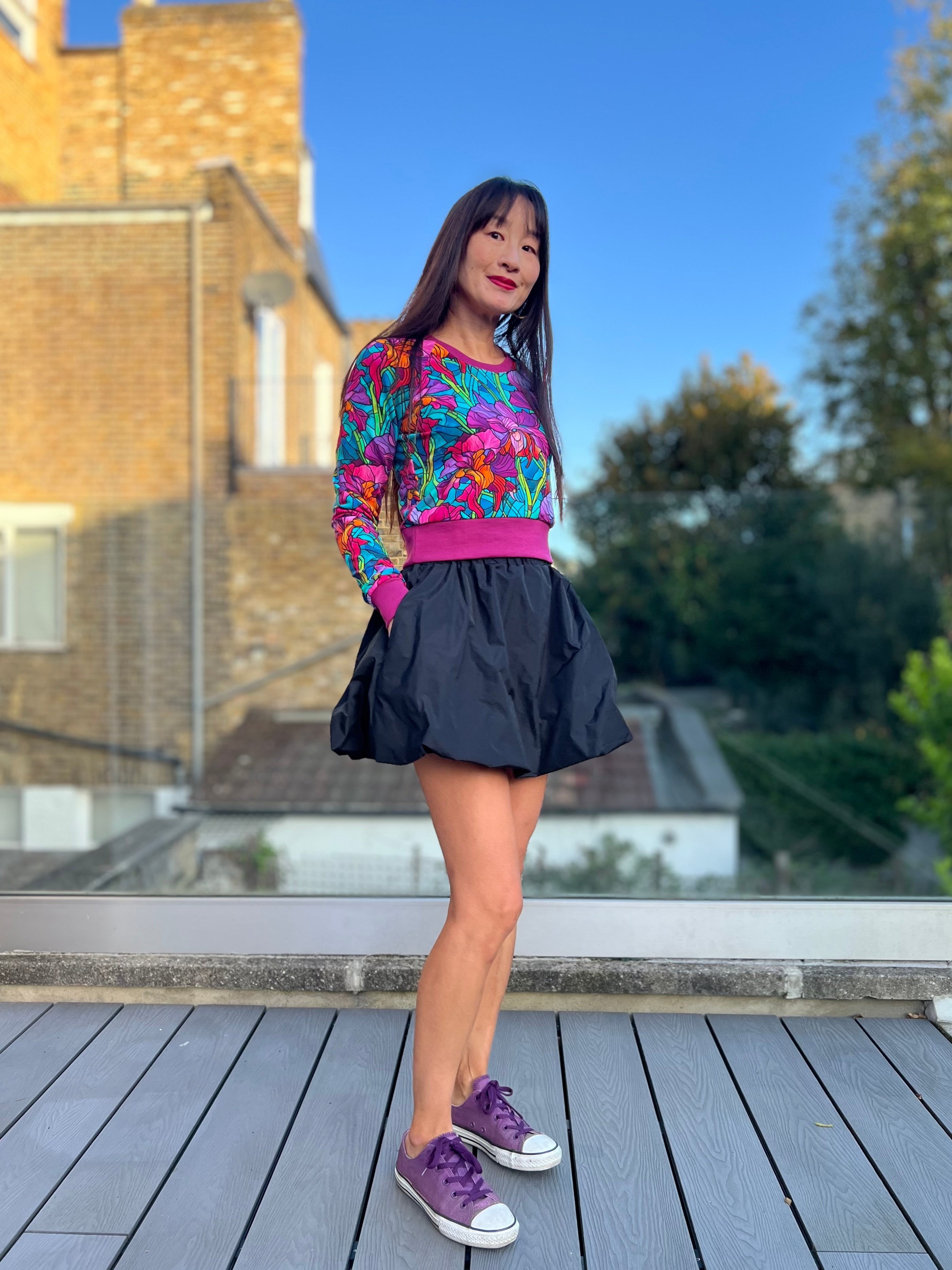 Woman wearing a colorful sweater and black taffeta bubble skirt on a wooden deck with a building and trees in the background. Model used the Sadie Bubble Skirt PDF sewing pattern to make the skirt.