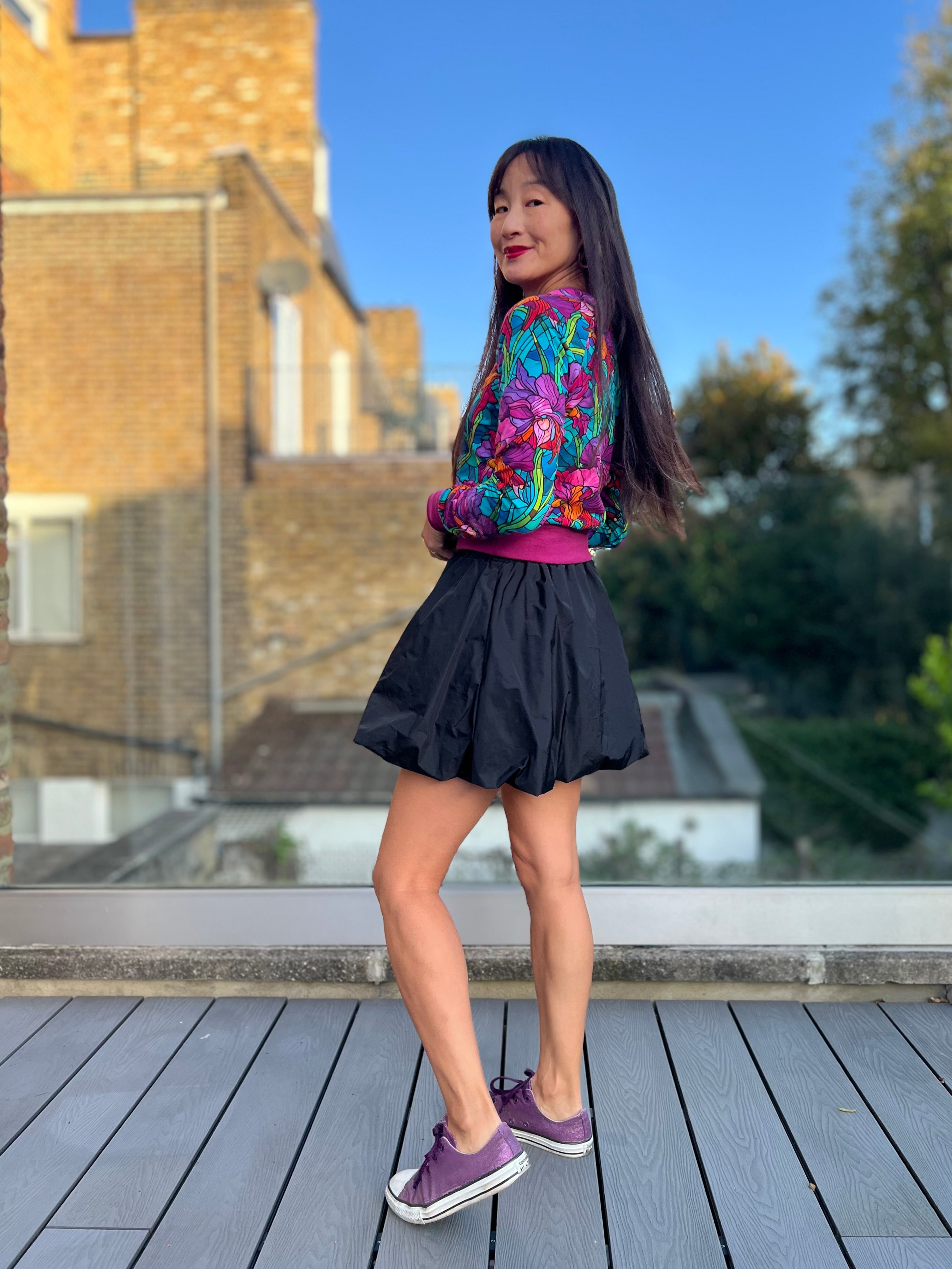 Woman in a colorful jacket and black mini skirt with pockets standing on a wooden deck with a building and trees in the background.