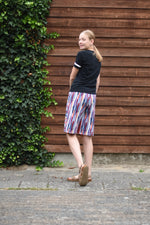 Person wearing a black t-shirt and colorful shorts standing against a wooden wall with greenery. Knee Length version of the  Caleb Athletic Sewing Pattern by Ellie and Mac Patterns.