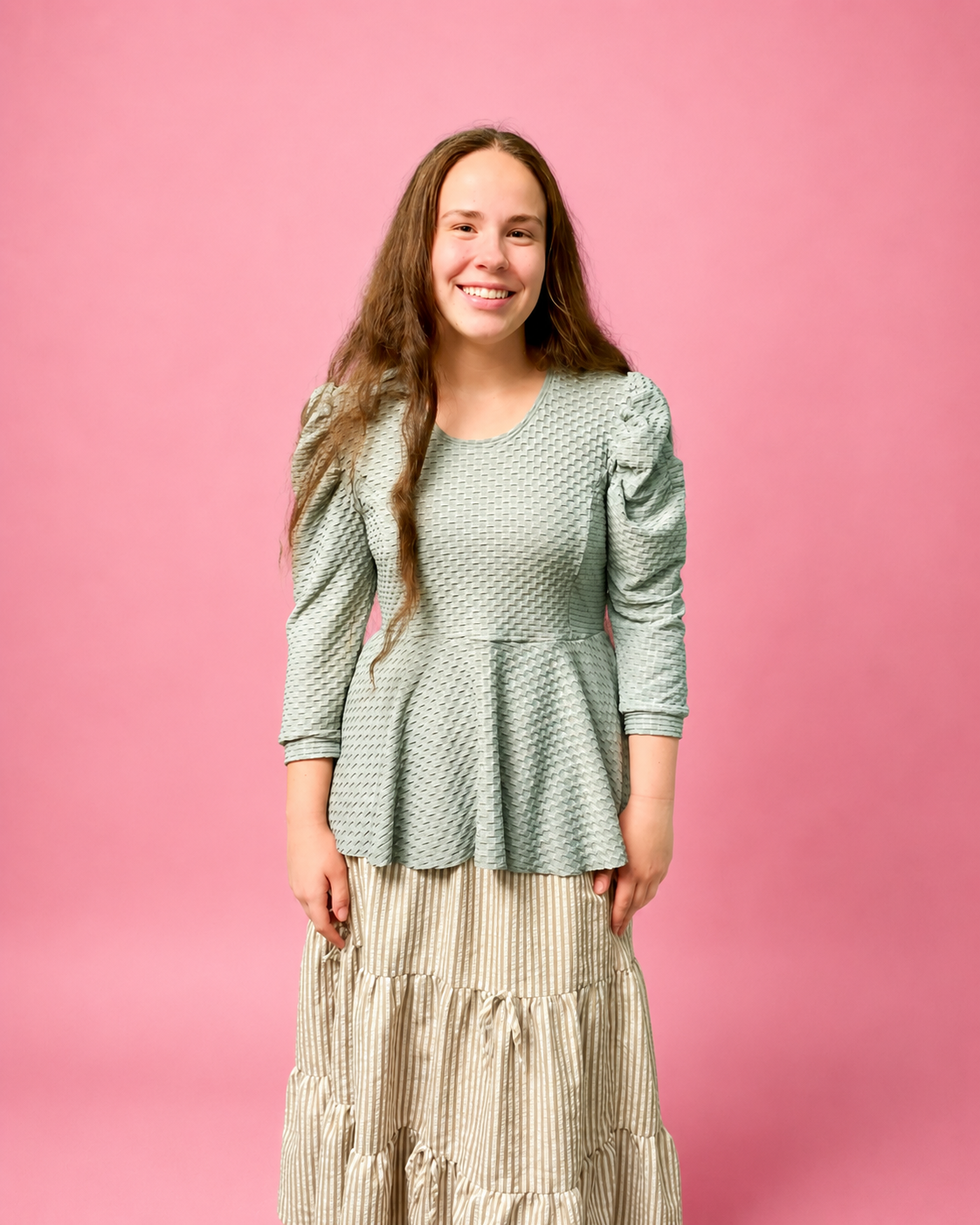 A young woman with long brown hair stands against a solid pink studio background, wearing a sage green textured long-sleeve peplum top and a beige and white vertically striped tiered skirt. The top is made using Ellie and Mac PDF sewing patterns. They include A4, Letter, A0, and Projector pattern files. They also include a full sew-along tutorial. 