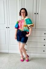 Woman wearing a colorful off-shoulder top and denim shorts standing in front of white cabinets. The blouse is made using Ellie and Mac PDF sewing patterns.