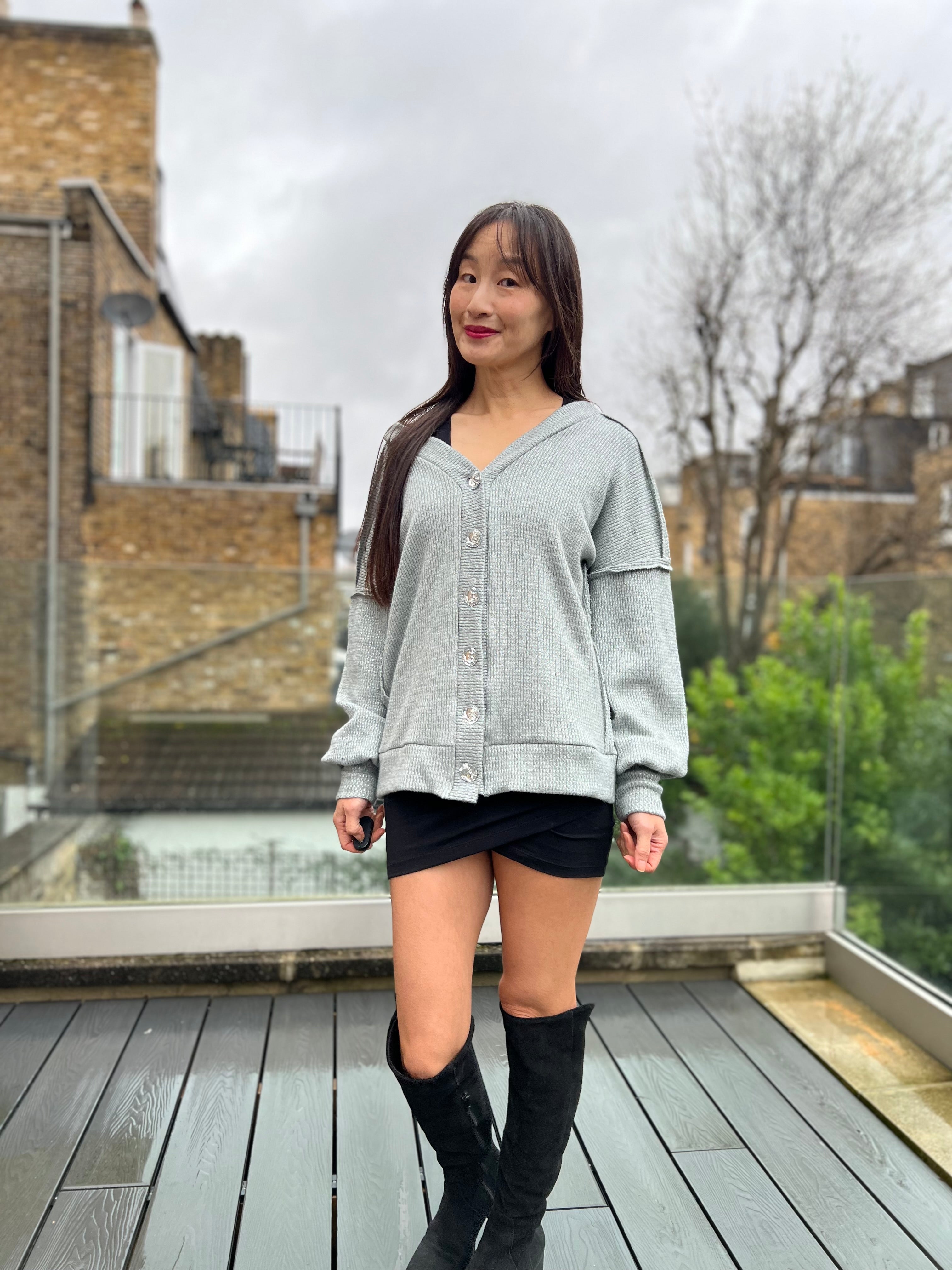 Woman wearing a gray oversized cardigan sweater and black dress standing on a wooden deck with a cityscape background.