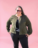 Person wearing a green jacket with zip and pockets and sunglasses against a pink background. Plus size sewing patterns xxs - 6xl size range. 