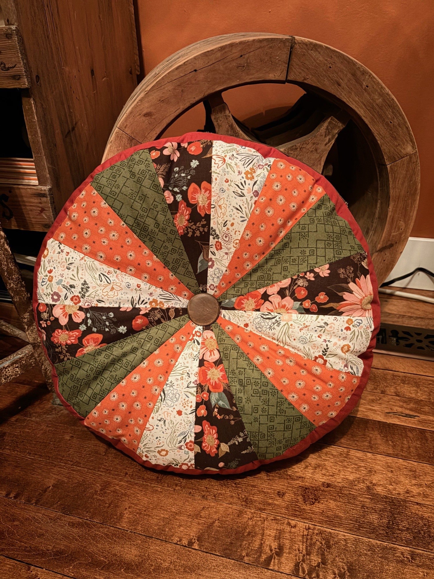 Patchwork wheel-shaped cushion on a wooden floor with a wooden wheel in the background. Easy home decor sewing patterns by Ellie and Mac.