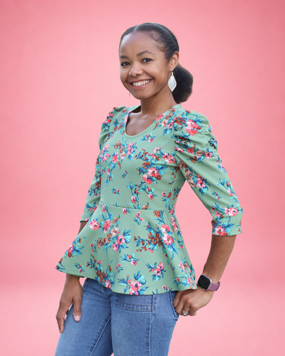 A woman modeling a sage green floral puff-sleeve peplum top with blue jeans, standing against a solid pink background. The top is made using Ellie and Mac PDF sewing patterns. They include A4, Letter, A0, and Projector pattern files. They also include a full sew-along tutorial. 