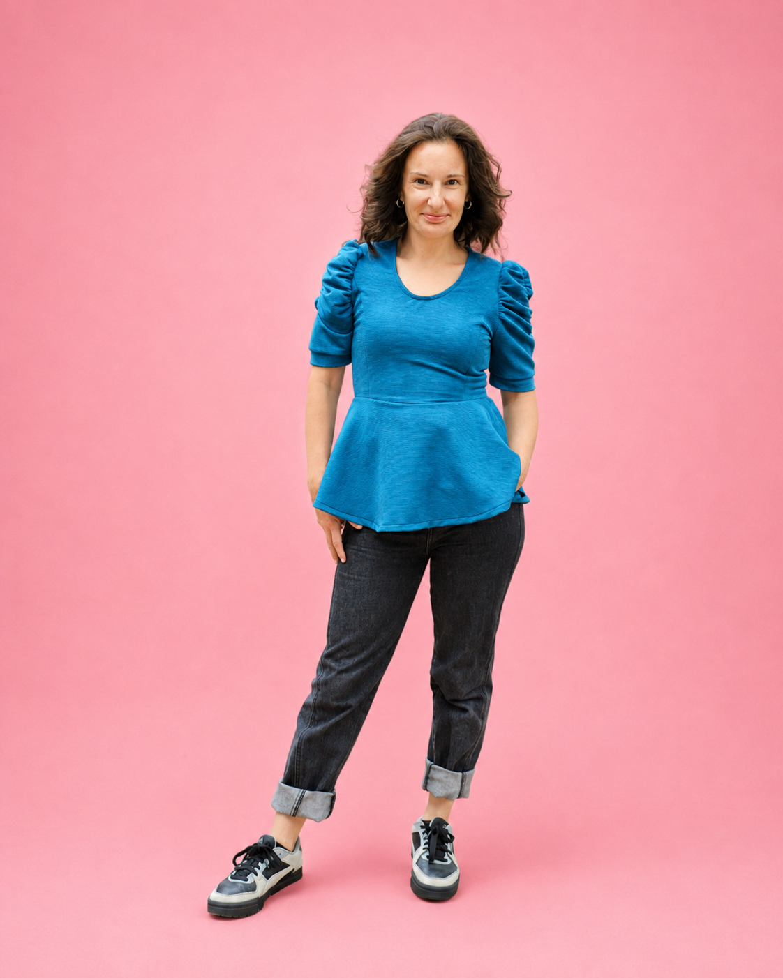 A woman modeling a bright blue short-sleeve peplum top, paired with dark gray cuffed jeans and black-and-white sneakers, standing against a solid pink studio background. The top is made using Ellie and Mac PDF sewing patterns. They include A4, Letter, A0, and Projector pattern files. They also include a full sew-along tutorial. 