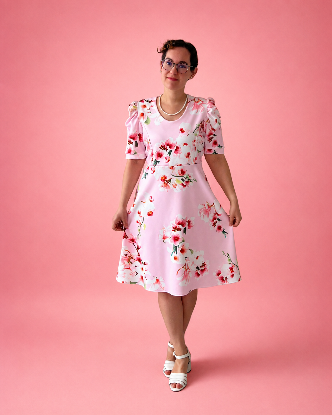 A woman wearing a light pink short-sleeve A-line dress with a red and white floral print, standing against a solid pink studio background. The dress is made using Ellie and Mac PDF sewing patterns. They include A4, Letter, A0, and Projector pattern files. They also include a full sew-along tutorial. 