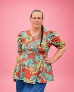 A woman modeling a short-sleeve peplum top with a tropical leaf print in green and teal on a coral-red background, paired with blue striped pants, set against a solid pink backdrop.