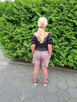 Girl standing next to a green bush on a paved path with blond hair, a black top, and cute shorts.