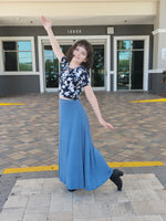 Woman in a floral top and blue skirt standing on a sidewalk with a building in the background