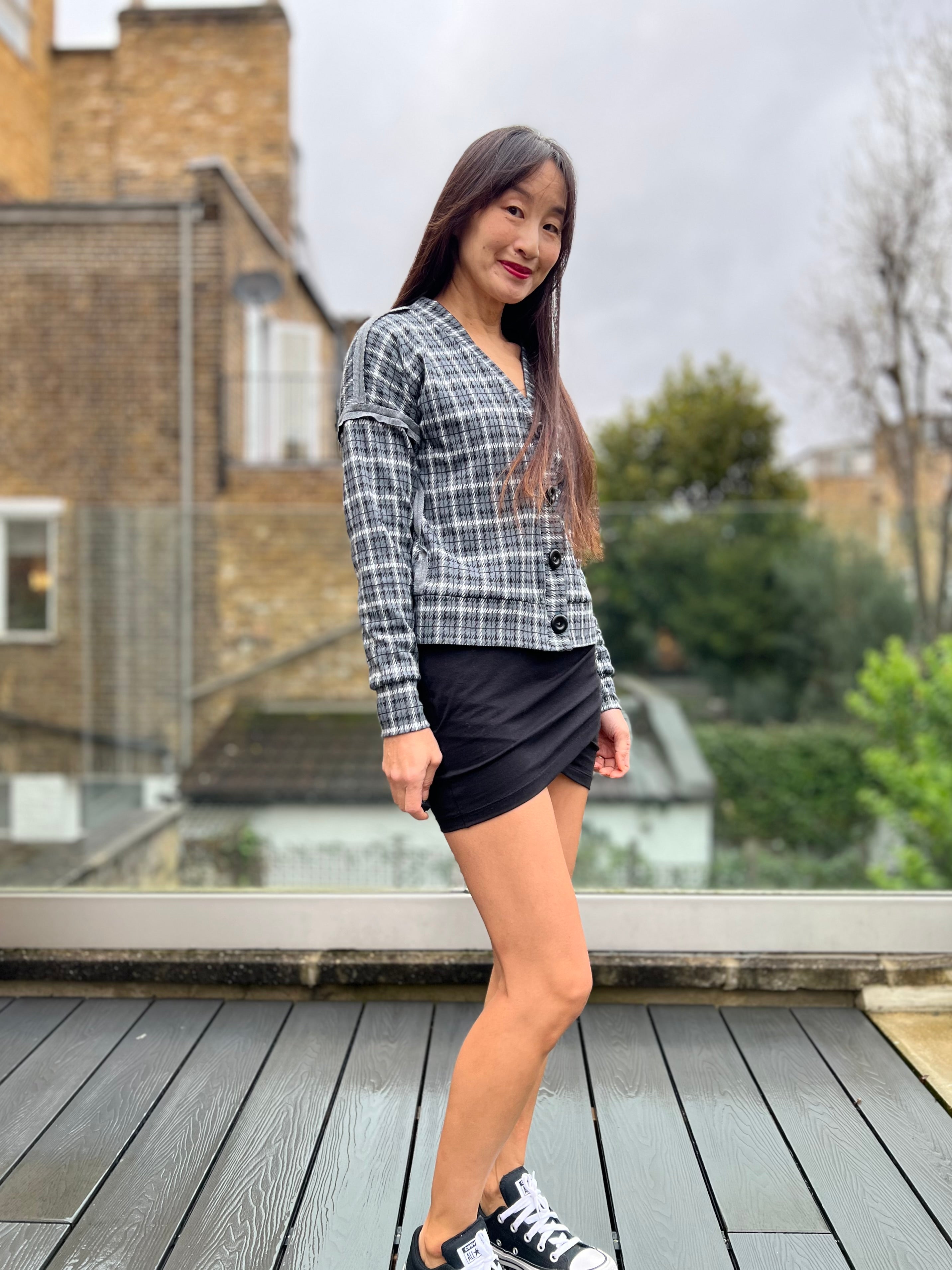 Woman standing on a wooden deck wearing a plaid cropped cardigan and black skirt.