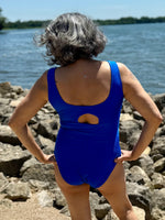 Person wearing a blue swimsuit standing on rocks by a body of water.
