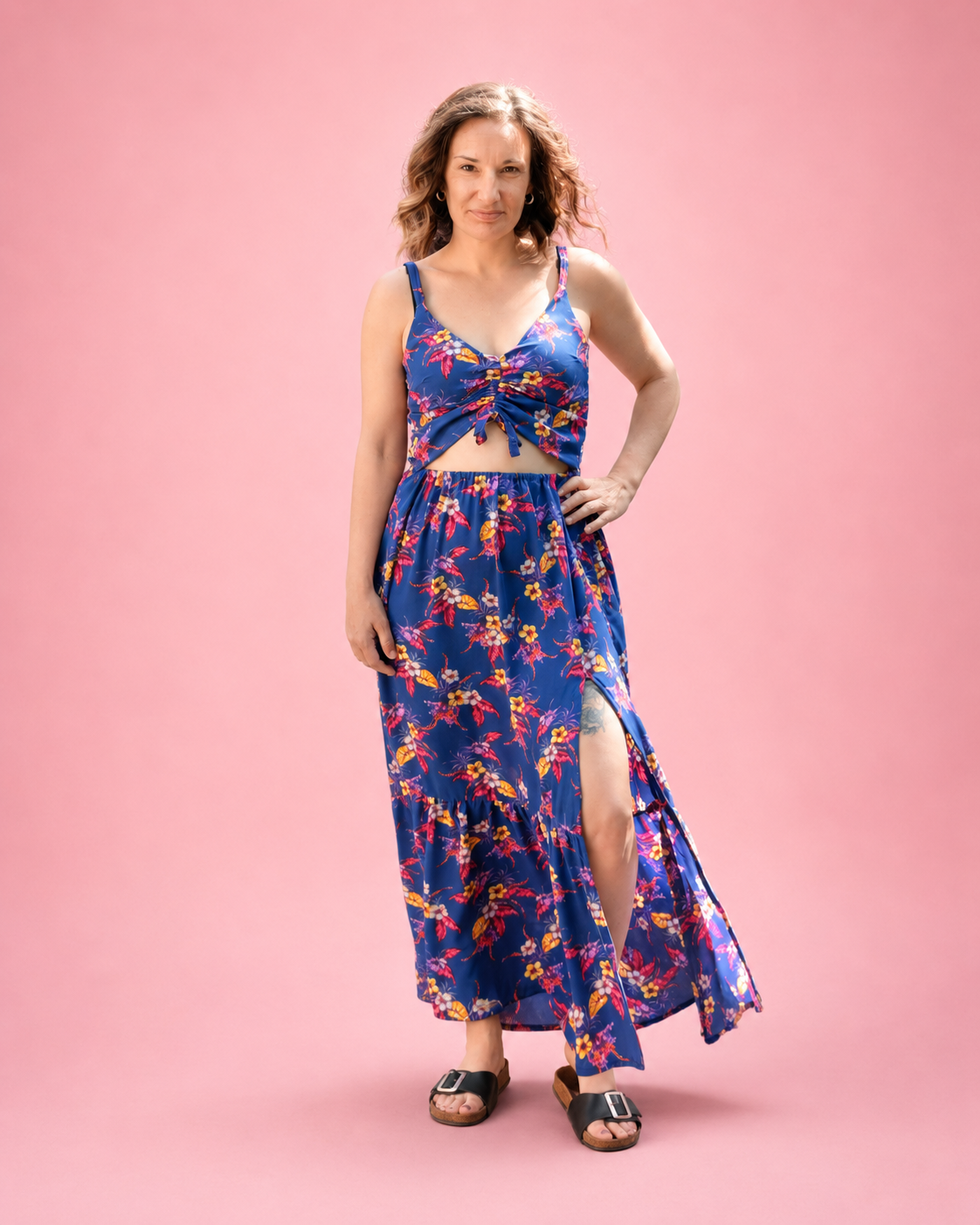 A woman modeling a woven blue floral maxi dress with a ruched tie-front bodice and a skirt with a high slit, standing against a solid pink studio background. Trendy sewing pattern by Ellie and Mac for woven fabrics. Suitable for beginners. Shirred back.