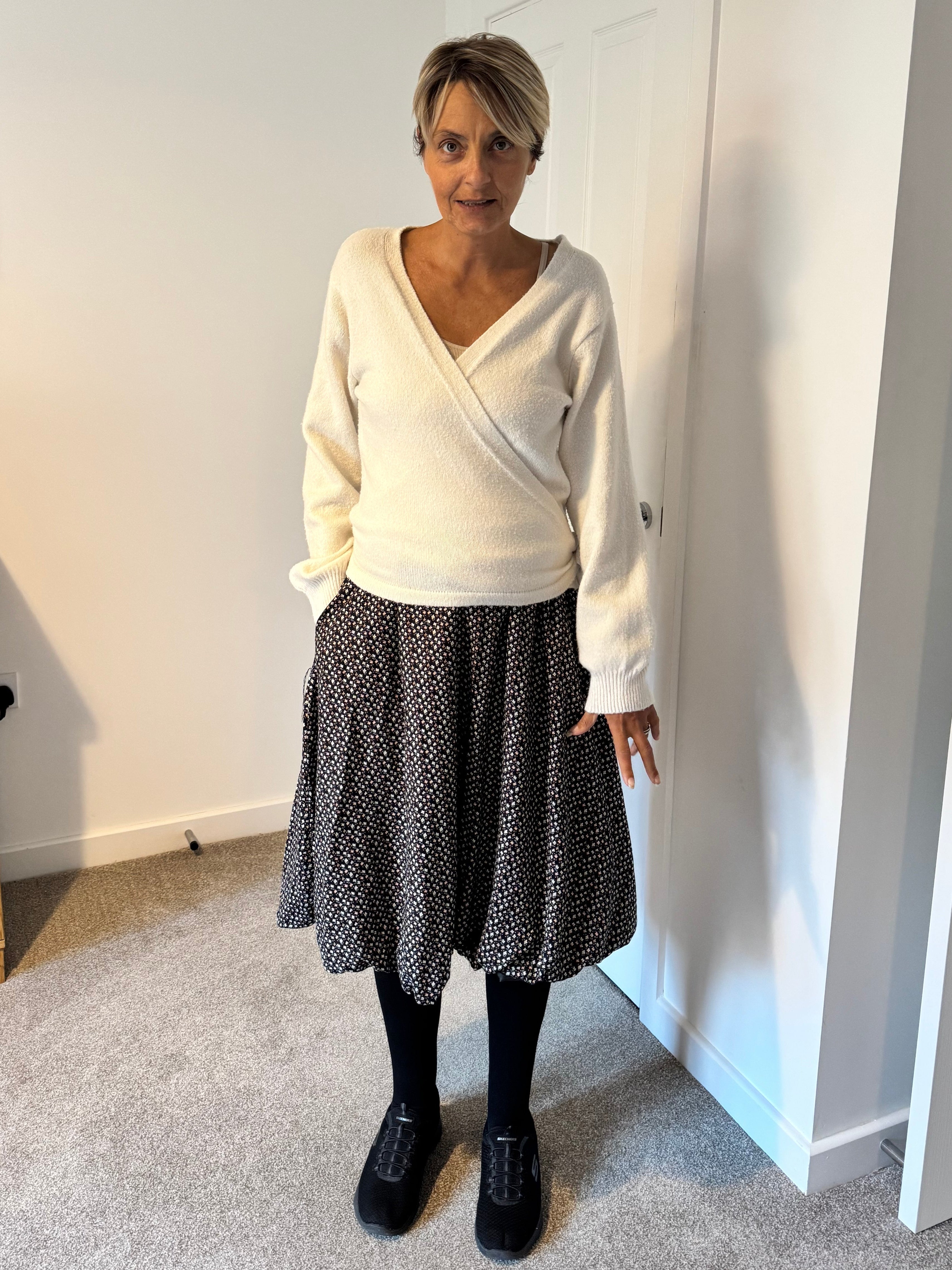 Person wearing a white top and black patterned skirt standing in a room with a door and wall decorations.