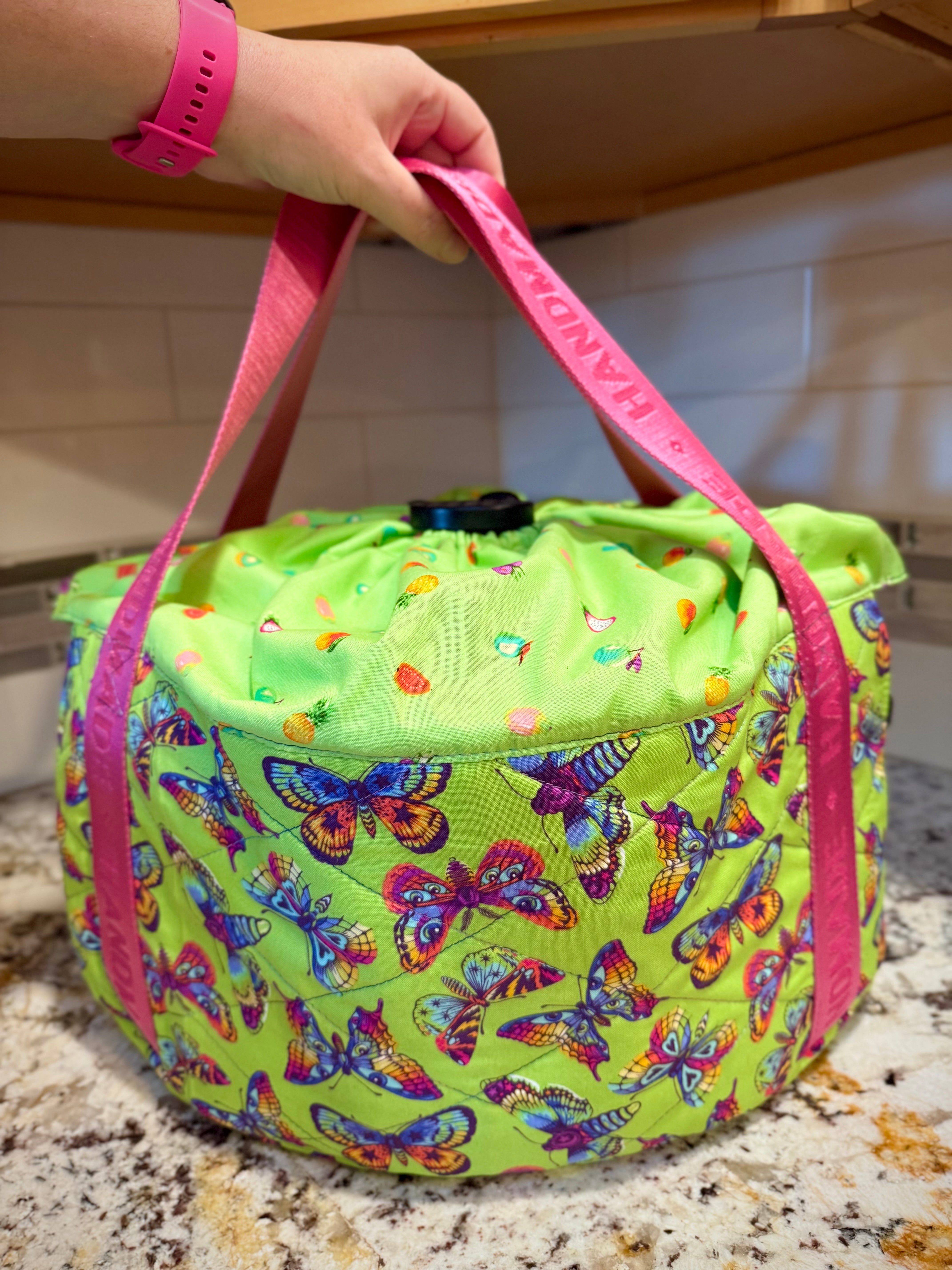 Green slow cooker carrier with butterfly pattern with hot pink straps, held by a hand on a kitchen counter. Made using the Ellie and Mac Warm and Cozy Slow Cooker Carrier Pattern.