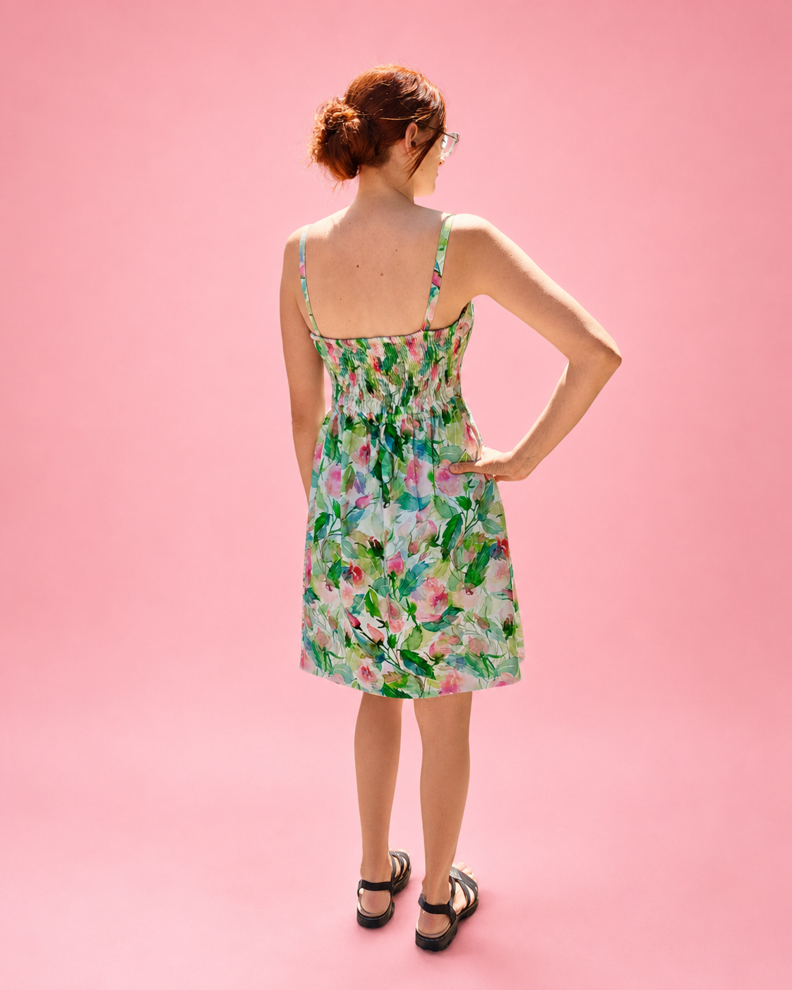 A woman wearing a sleeveless floral dress is photographed from the back against a solid pink studio background. The dress has a green, pink, and white watercolor floral pattern with thin straps and a shirred back bodice. Woven sewing pattern by Ellie and Mac.