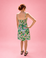 A woman wearing a sleeveless floral dress is photographed from the back against a solid pink studio background. The dress has a green, pink, and white watercolor floral pattern with thin straps and a shirred back bodice. Woven sewing pattern by Ellie and Mac.