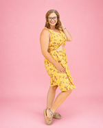 A woman modeling a yellow sleeveless dress with a small floral print, featuring a  front ruched cutout, posed against a solid pink background. Perfect woven sewing patterns.