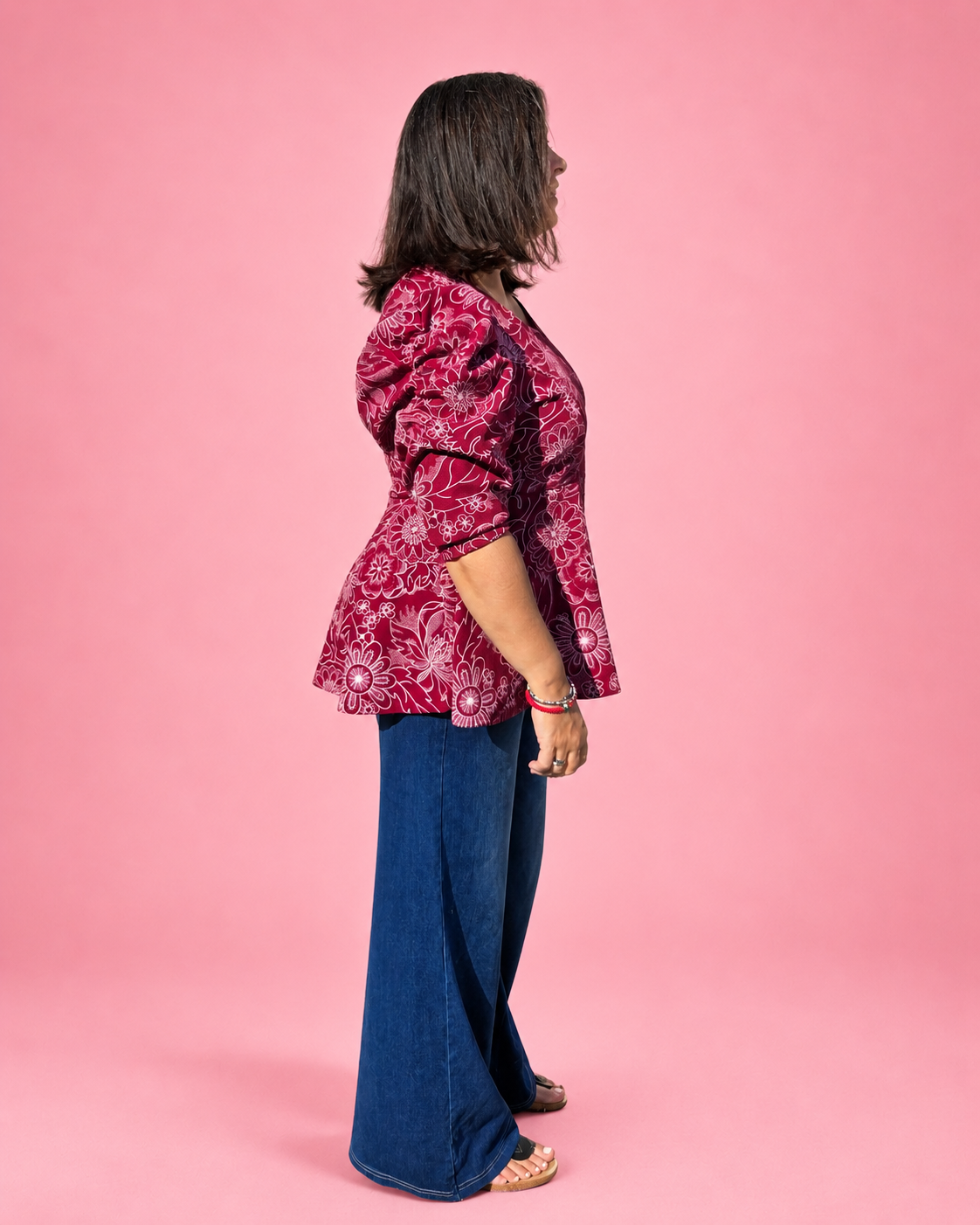 A woman shown in side profile wearing a dark red and white paisley patterned peplum blouse with three-quarter puffed sleeves, paired with wide-leg blue jeans, standing against a solid pink studio background. The top is made using Ellie and Mac PDF sewing patterns. They include A4, Letter, A0, and Projector pattern files. They also include a full sew-along tutorial. 