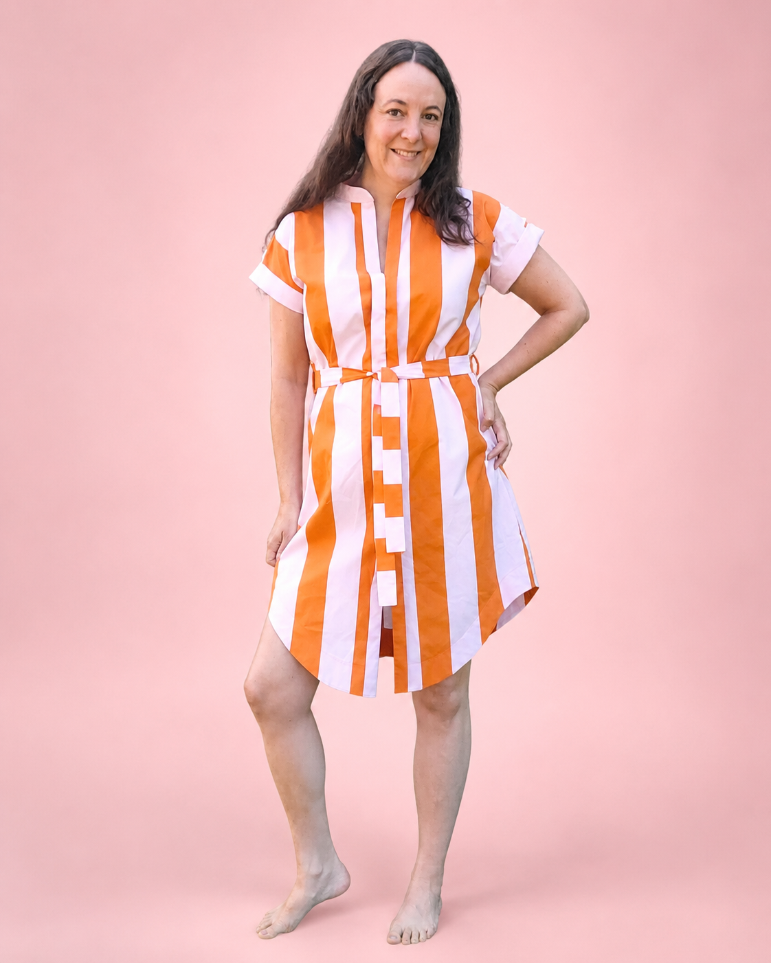 A woman modeling a short-sleeve button-front dress she made with bold vertical orange and white stripes, standing barefoot against a solid pink studio background.