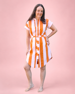 A woman modeling a short-sleeve button-front dress she made with bold vertical orange and white stripes, standing barefoot against a solid pink studio background.