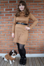 Woman in a brown dress standing next to a dog in front of a brick wall. Dog is standing next to her wearing a matching sweater.