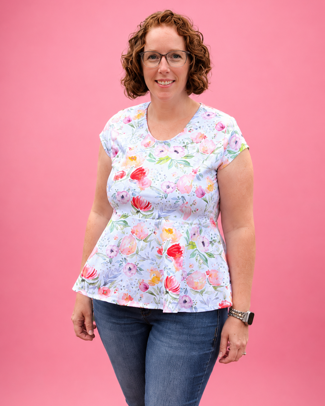 A woman with curly brown hair and glasses wearing a light blue short-sleeve peplum top with a colorful floral print, paired with blue jeans, standing against a solid pink background. The top is made using Ellie and Mac PDF sewing patterns. They include A4, Letter, A0, and Projector pattern files. They also include a full sew-along tutorial. 