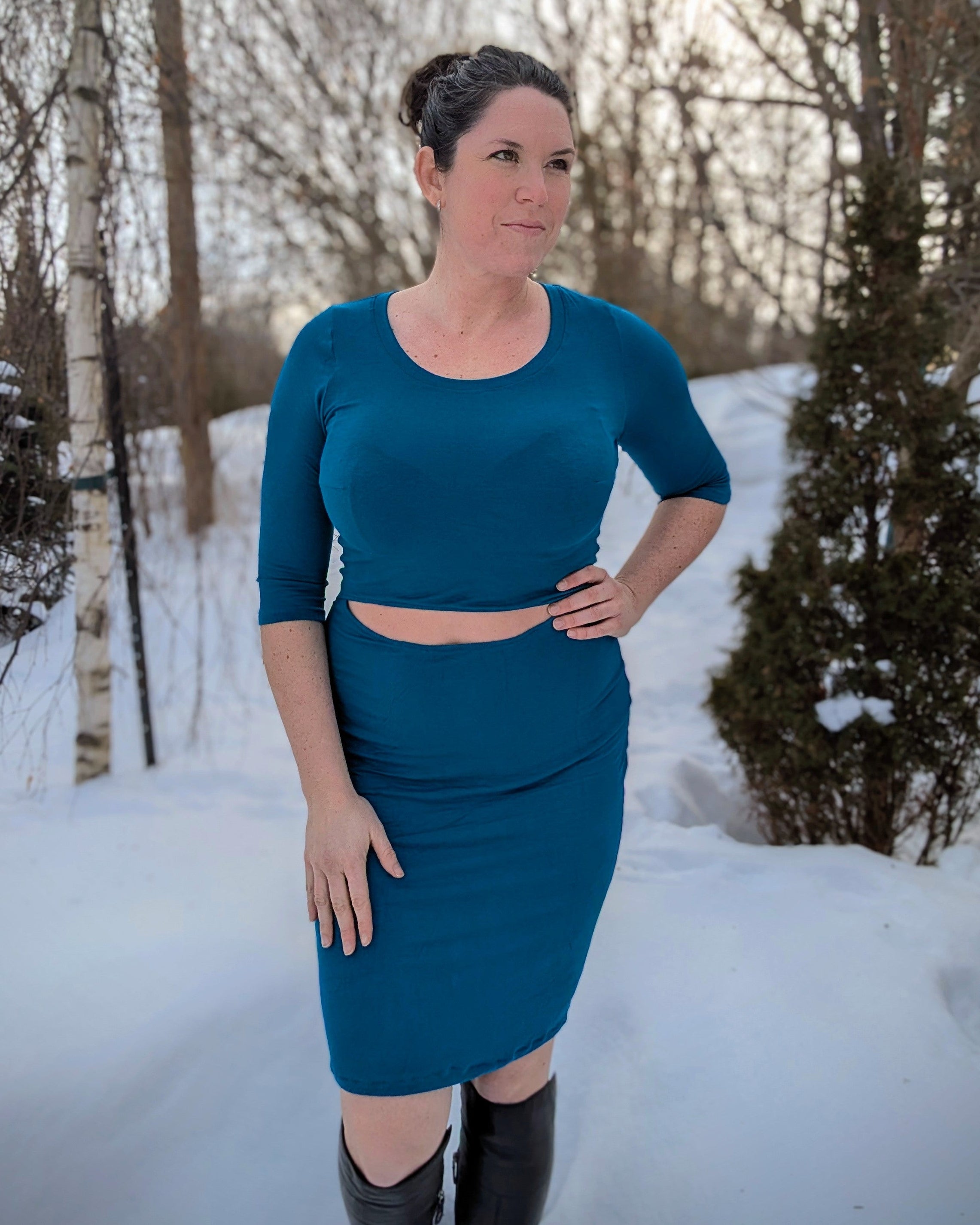 Woman in a teal fitted dress with front cutout standing in a snowy landscape with trees.