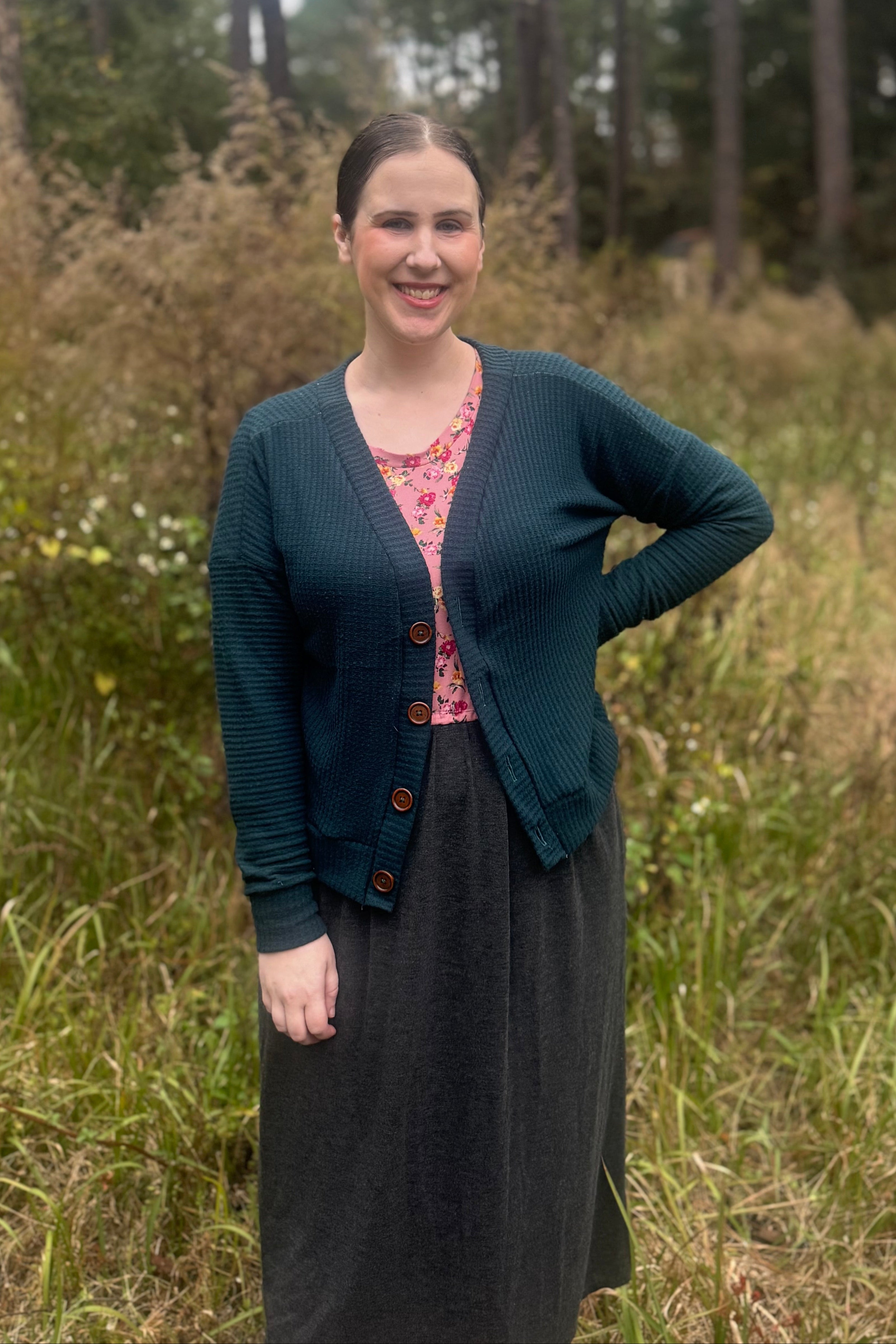 Woman standing in a forested area wearing a dark green cardigan and long dark skirt.