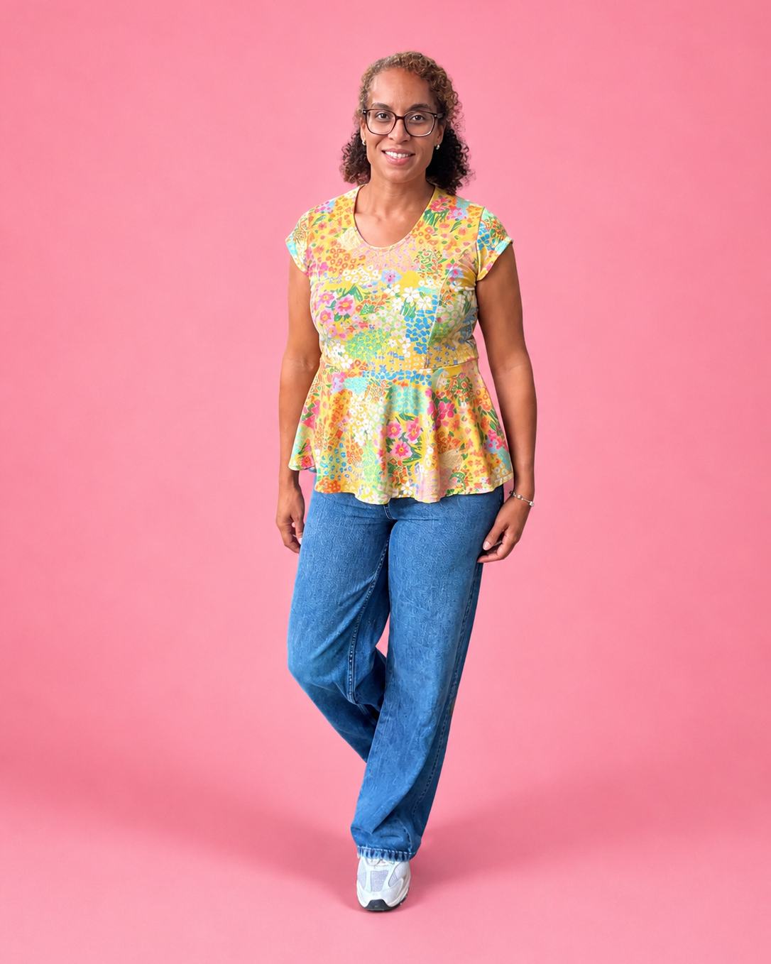 A woman wearing a colorful floral short-sleeve peplum top and blue jeans stands against a solid pink background. The top is made using Ellie and Mac PDF sewing patterns. They include A4, Letter, A0, and Projector pattern files. They also include a full sew-along tutorial. Easy sewing for beginners.