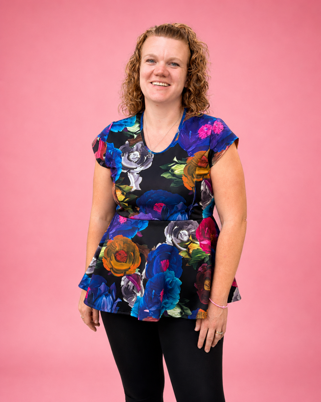 A woman with curly light brown hair wearing a short-sleeve peplum top with a vibrant multicolor floral print on a black background, paired with black leggings, standing against a solid pink studio backdrop. The top is made using Ellie and Mac PDF sewing patterns. They include A4, Letter, A0, and Projector pattern files. They also include a full sew-along tutorial. 
