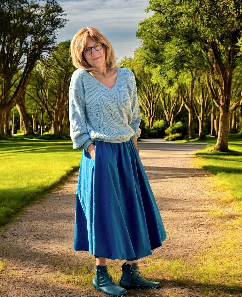 Woman in a blue sweater and ankle bubble skirt standing on a path with trees in the background.