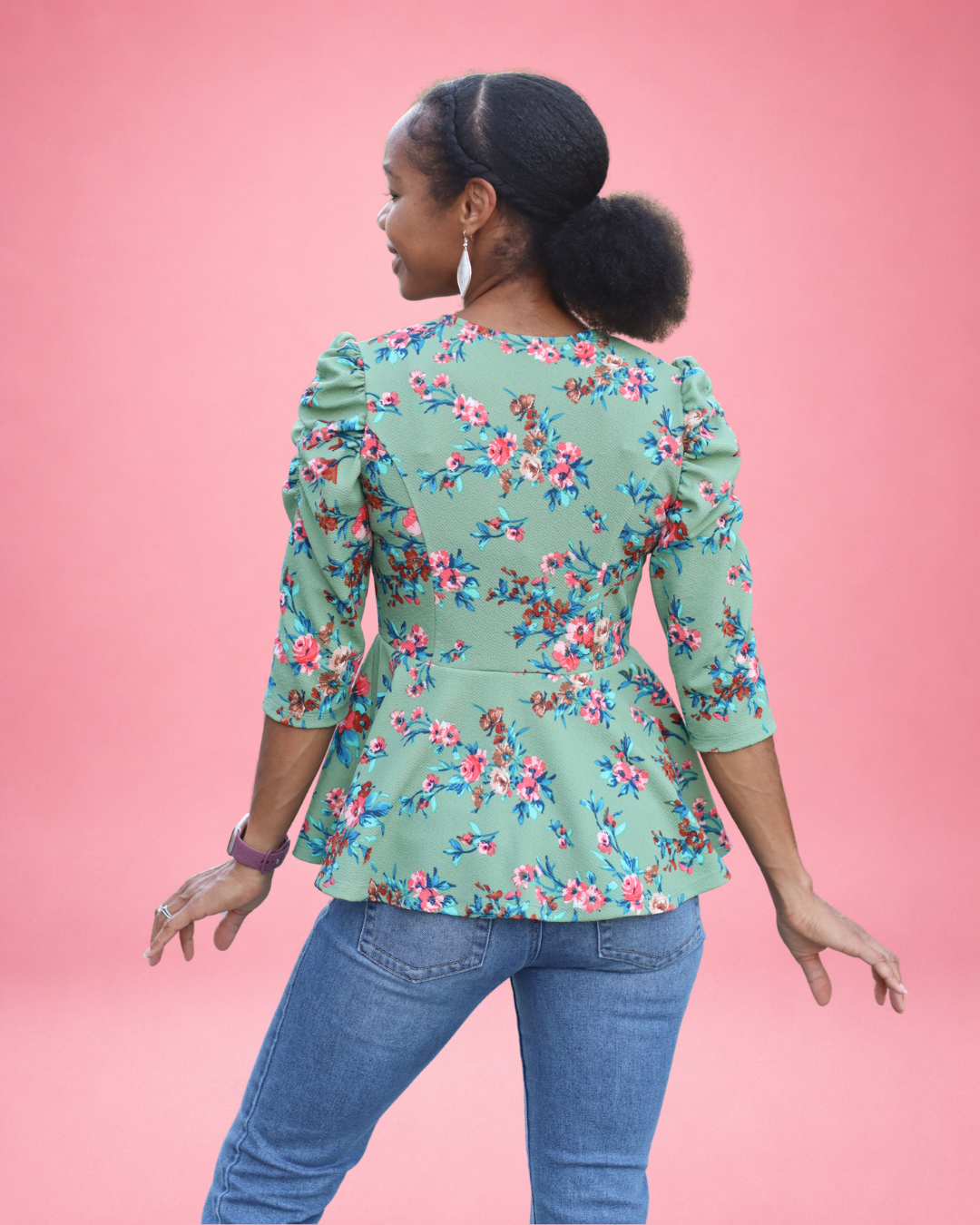 A woman is shown from the back wearing a sage green peplum top with a pink, blue, and red floral pattern, paired with blue jeans, set against a solid pink background. The top is made using Ellie and Mac PDF sewing patterns. They include A4, Letter, A0, and Projector pattern files. They also include a full sew-along tutorial. 