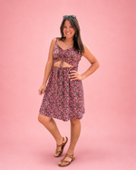 A woman modeling a sleeveless floral mini dress with a tie-front bodice that creates a trendy cutout, paired with brown sandals, against a solid pink studio background. Ellie and Mac Adeline Ruched Front Sundress sewing pattern for woven fabrics.