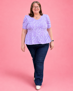 A woman wearing a light purple lace short-sleeve peplum top and dark blue jeans, standing against a solid pink studio background.