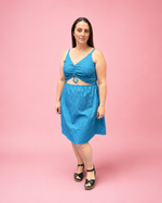 A woman modeling a bright blue textured two-piece set consisting of a halter crop top with a front cutout and a matching knee-length skirt, set against a solid pink studio background.